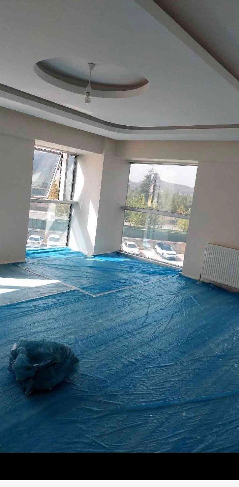 Tepsi tavanlı salon, dağ ve ağaç manzaralı pencereler, zemine serilmiş mavi koruma örtüsü