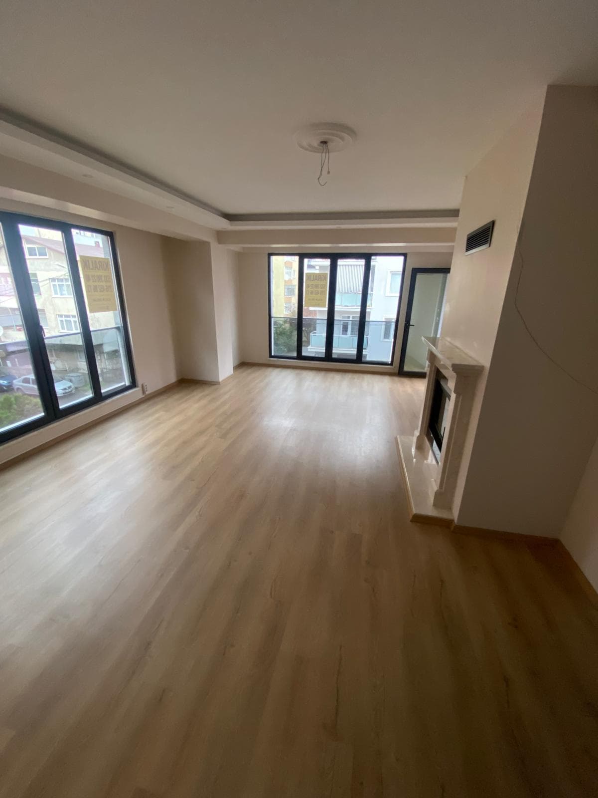 İkinci bir mermer/traverten şömine detaylı boş salon, bal rengi laminat zemin, pencerede 'KİRALIK' tabelası iki kez görünüyor