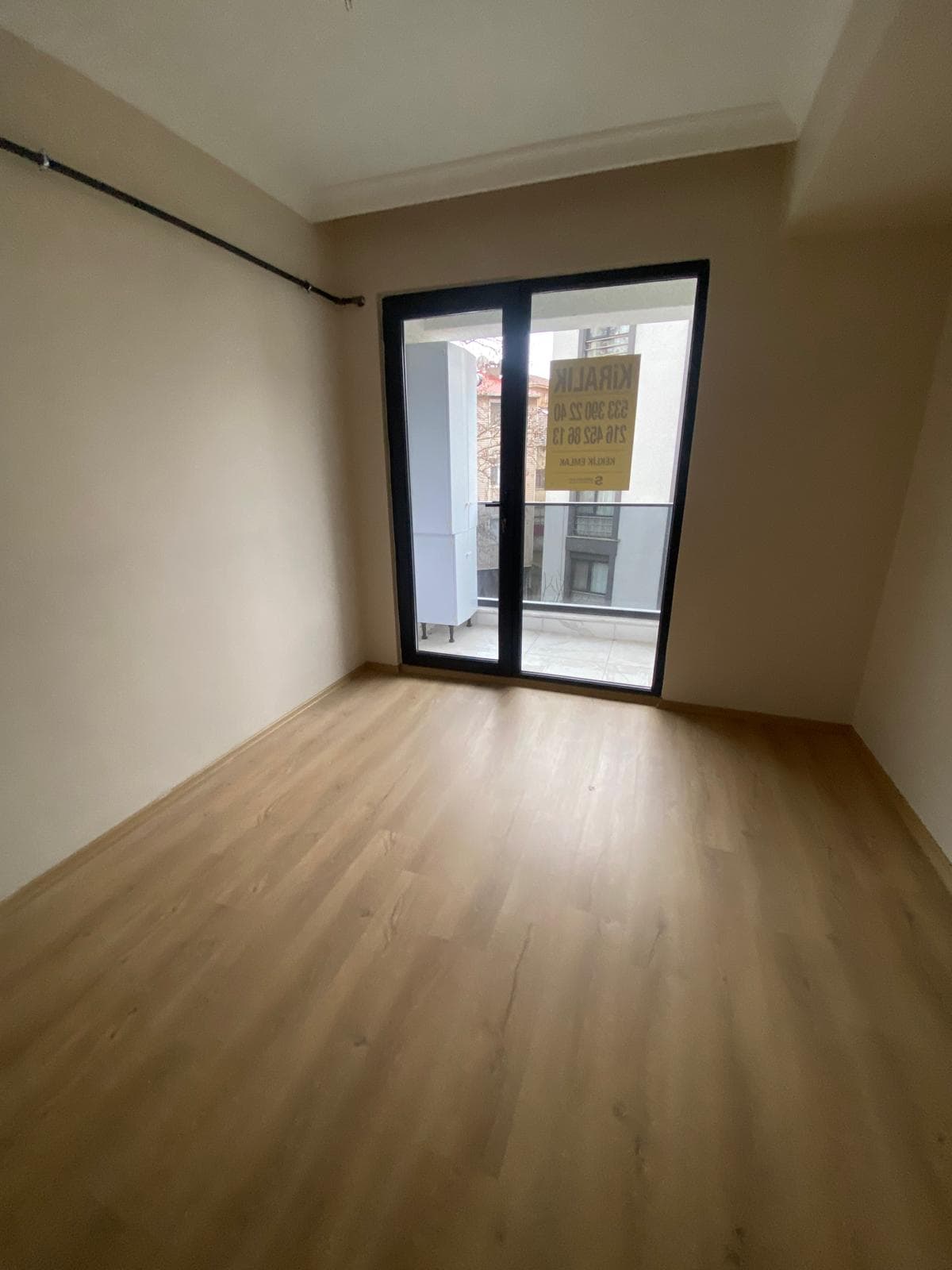 Boş oda, bej duvarlar, açık kahverengi laminat zemin, balkon kapısı camında 'KİRALIK' tabelası yansıması